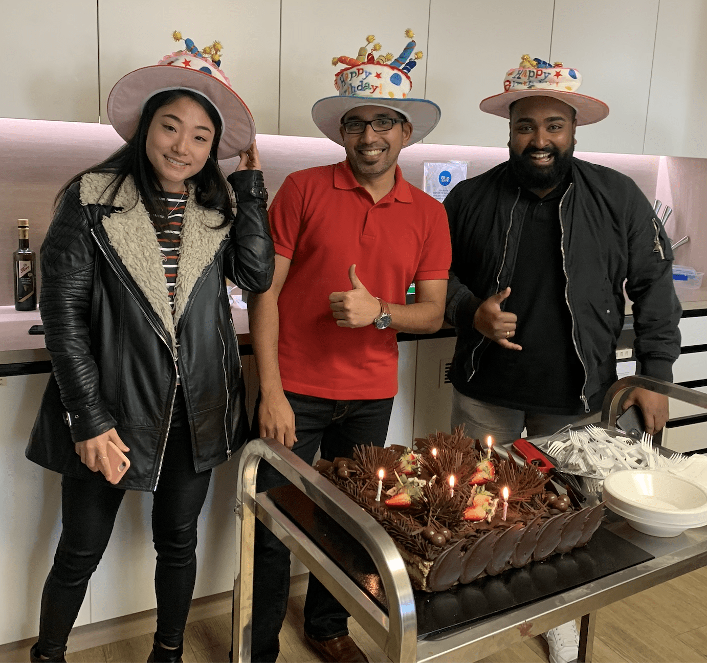 Three happy employees standing in front of a birthday cake at a company event